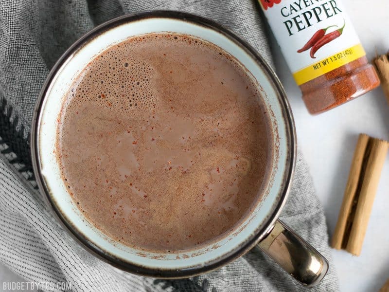 Overhead view of a mug full of Spicy Hot Cocoa with a bottle of cayenne and cinnamon sticks on the side