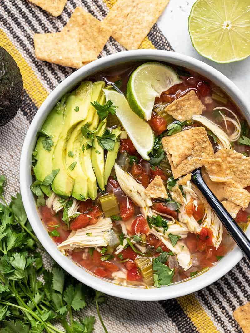 Close up overhead view of a bowl of Chicken and Lime Soup with avocado slices, lime wedges, and tortilla chips.