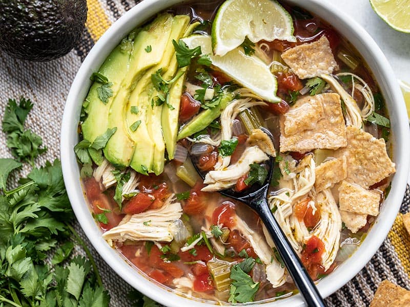 Close up of a bowl of Chicken and Lime Soup with garnishes and a black spoon in the middle