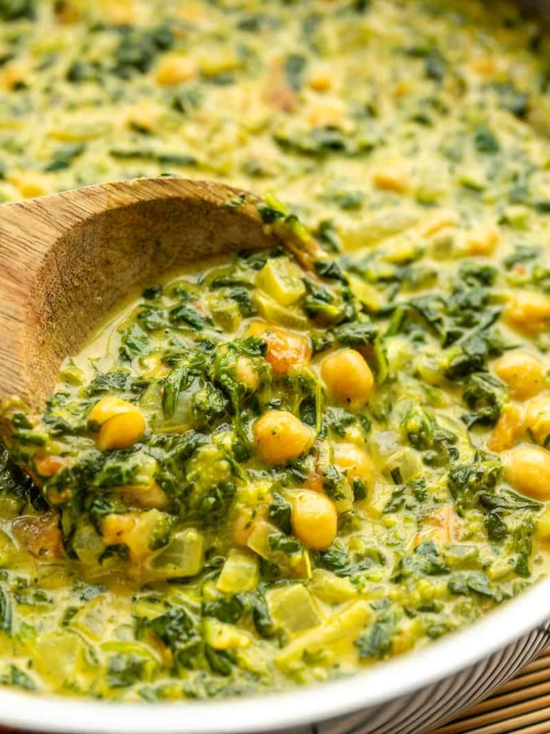 Close up of a wooden spoon scooping curried chickpeas and spinach out of a skillet