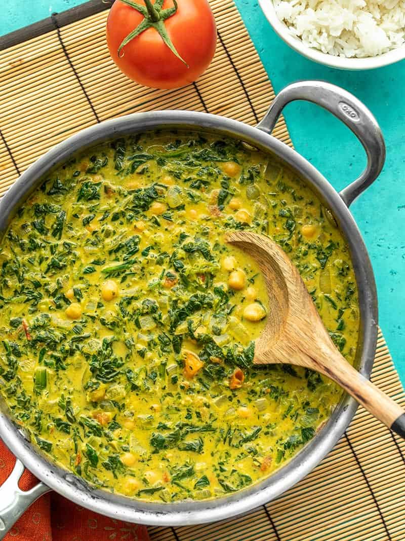 A skillet full of chana saag with a wooden spoon, on a bamboo mat next to a tomato and bowl of rice