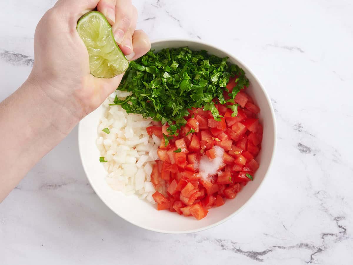 Ingredients for pico de gallo in a bowl with a hand squeeze a lime into it.