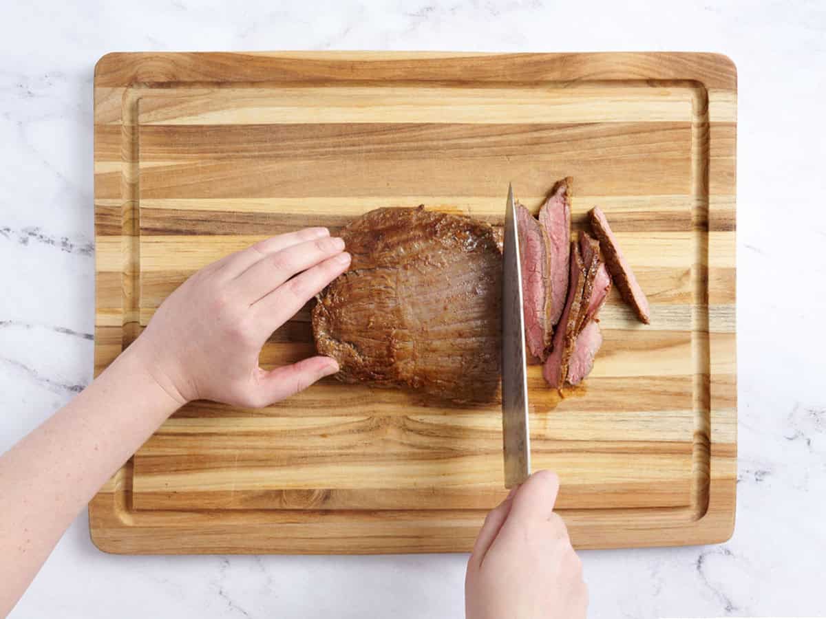 Hands using a knife to slice a cooked flank steak on a wooden cutting board.