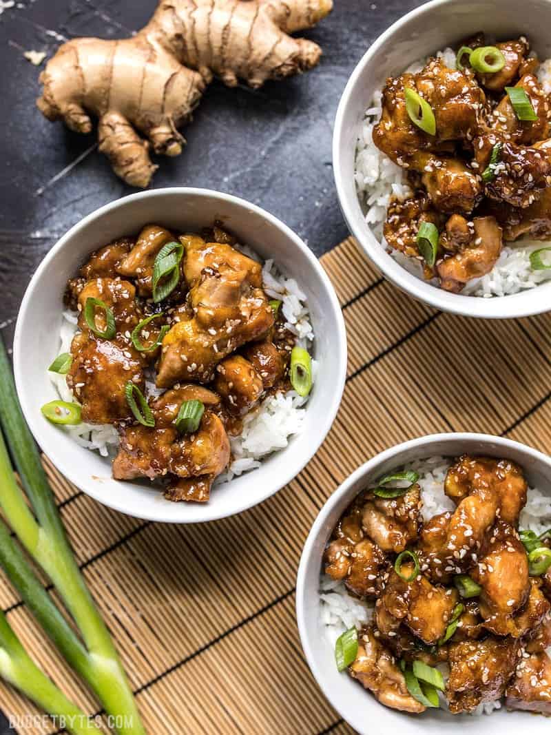 Three bowls of easy sesame chicken on a bamboo mat with ginger and green onion next to the bowls. 