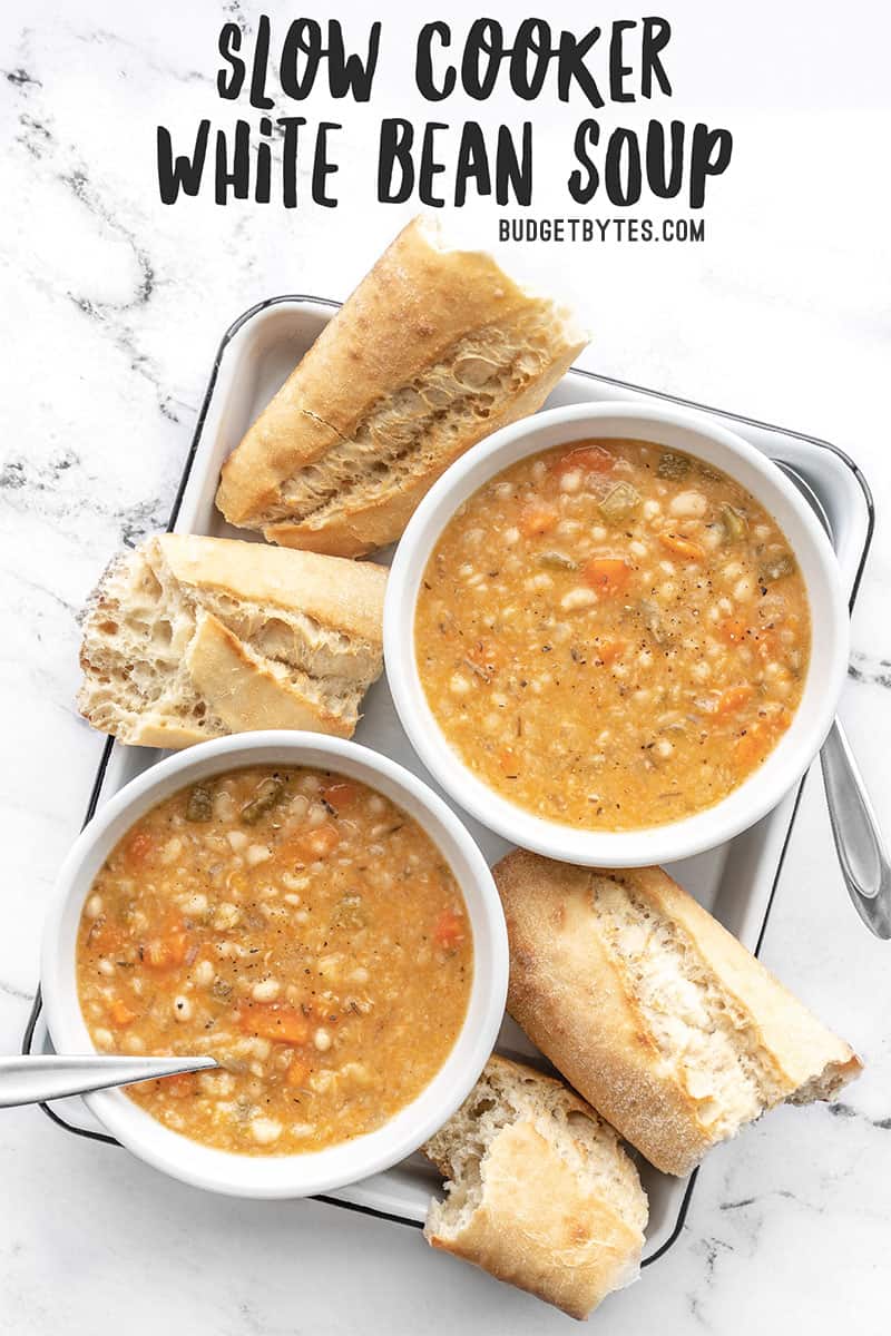 Two bowls of slow cooker white bean soup on a white tray with baguette pieces, title text at the top