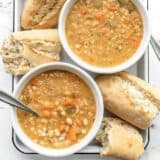 Two bowls of white bean soup on a white tray with torn baguette pieces on the side