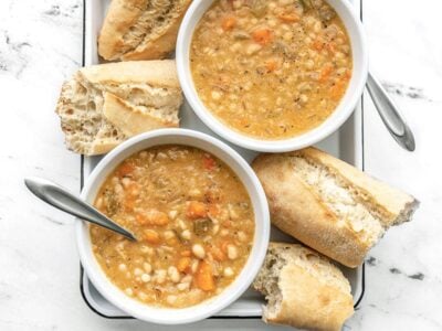 Two bowls of white bean soup on a white tray with torn baguette pieces on the side