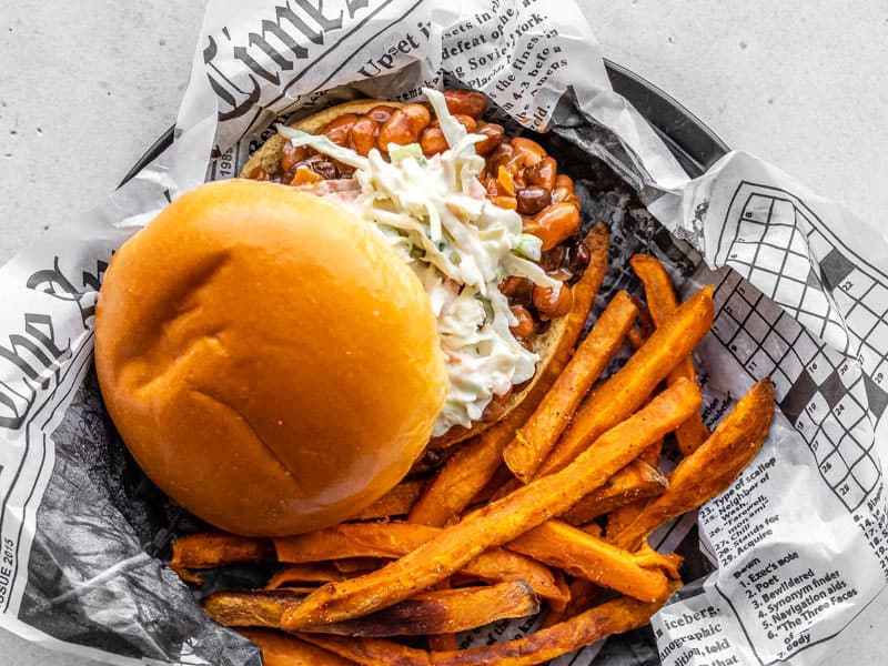 These BBQ Bean Sliders topped with a homemade creamy coleslaw are a quick dinner fix when you're craving BBQ and don't have time for slow cooking meat! BudgetBytes.com A plate covered in newsprint with a BBQ Bean Slider and some sweet potato fries