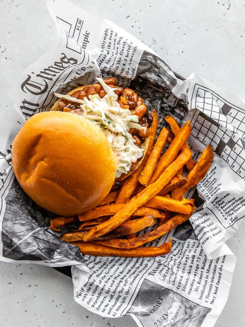 These BBQ Bean Sliders topped with a homemade creamy coleslaw are a quick dinner fix when you're craving BBQ and don't have time for slow cooking meat! BudgetBytes.com Overhead view of a plate covered with newsprint with one BBQ Bean Slider and some sweet potato fries