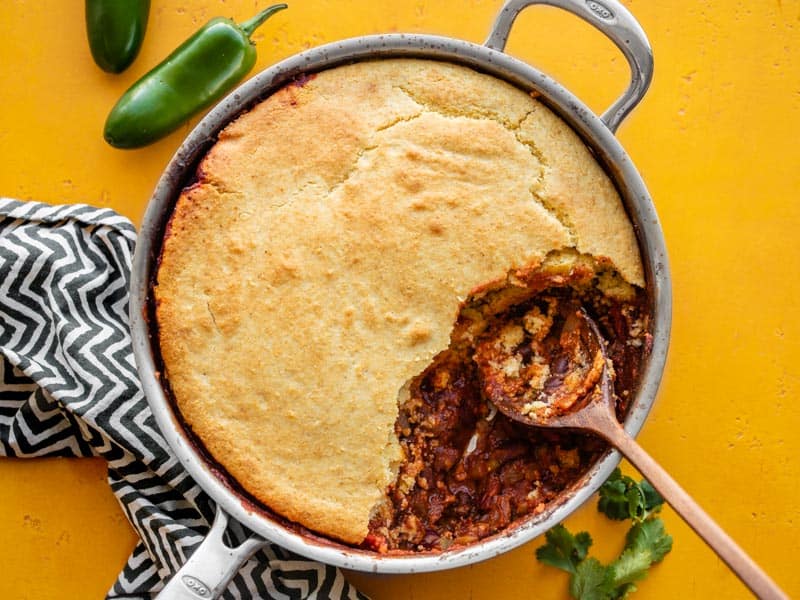 Chili Cornbread Skillet in the skillet with some scooped out, on a yellow surface with black and white striped napkin