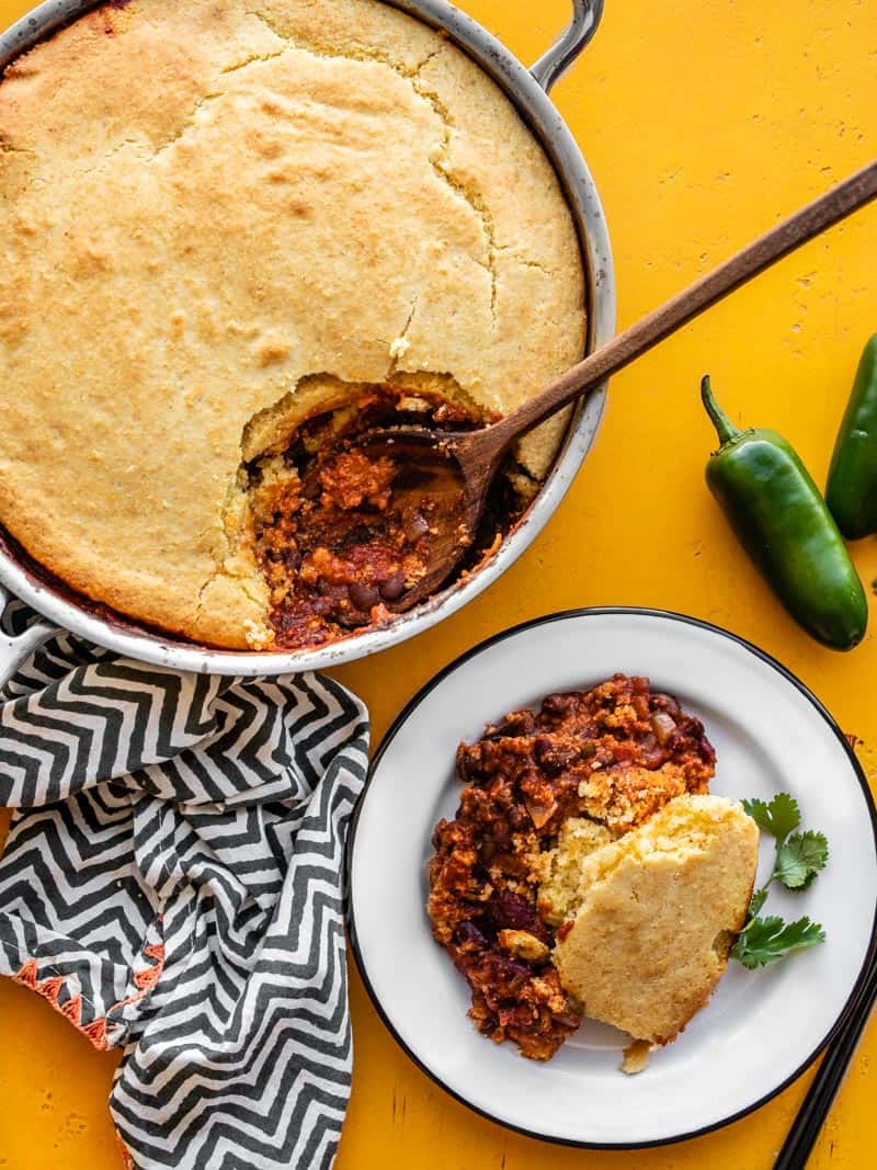 Chili cornbread skillet in the skillet with wooden spoon, a serving scooped out onto a plate on the side.