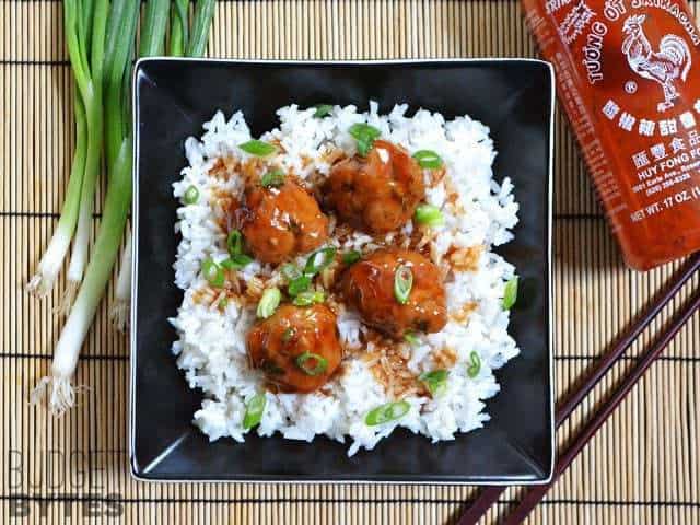 Top view of a bowl of Turkey Sriracha Meatballs over white rice with chopsticks and a bottle of Siracha on the side 