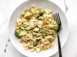 A bowl full of creamy pesto pasta with chicken and broccoli, a black fork on the side