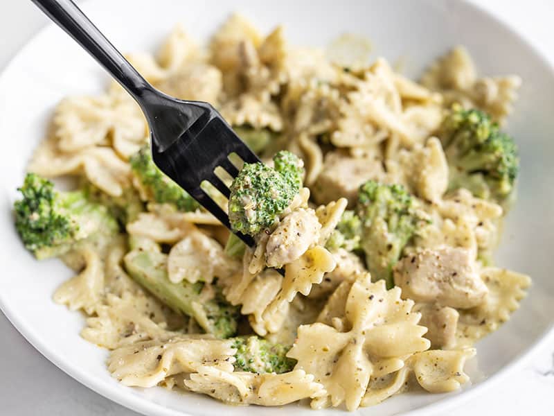 Front view of a bowl of creamy pesto pasta with chicken and broccoli, a fork lifting one bite