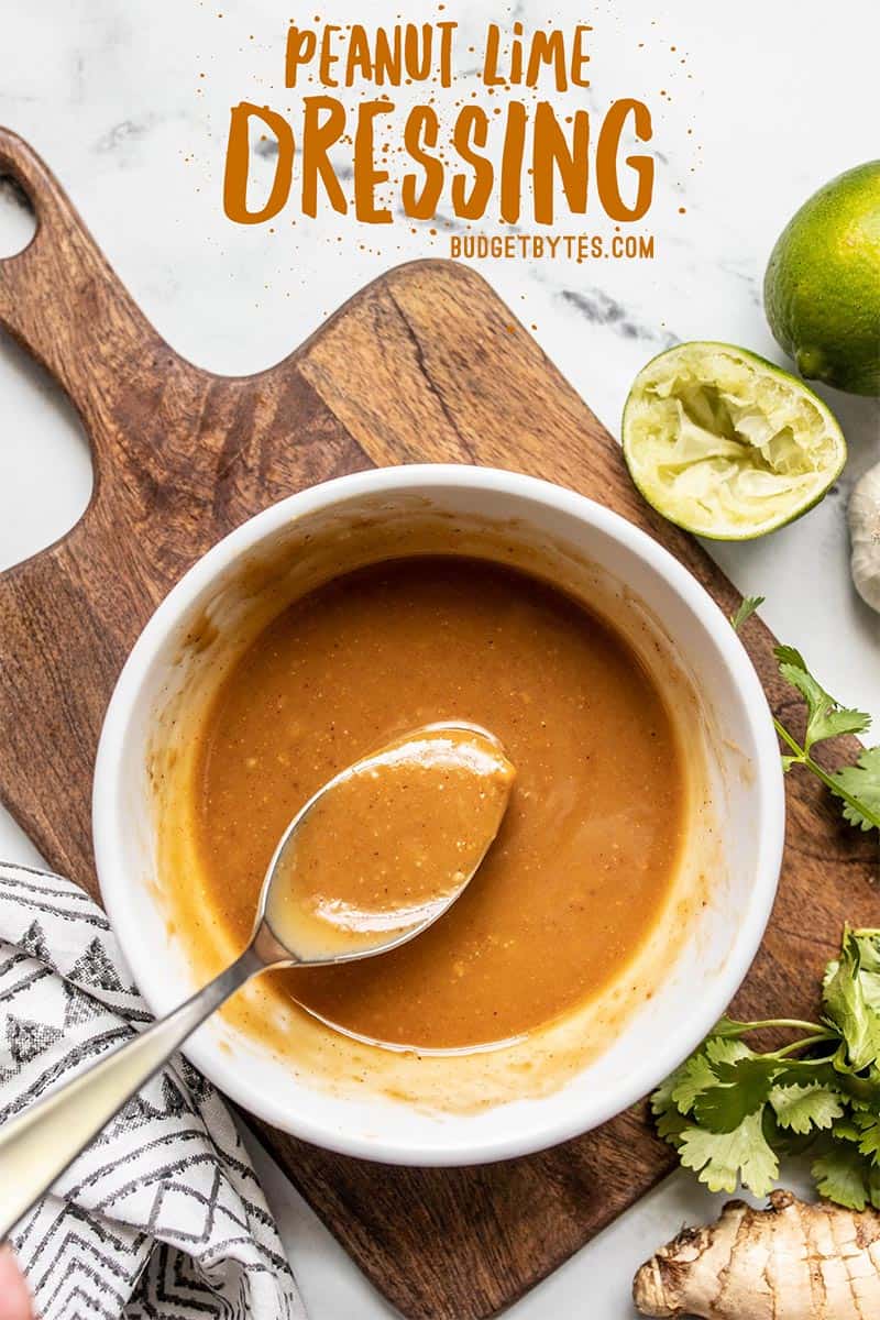 A bowl of peanut lime dressing on a cutting board with a spoon in the side, limes and cilantro on the side.