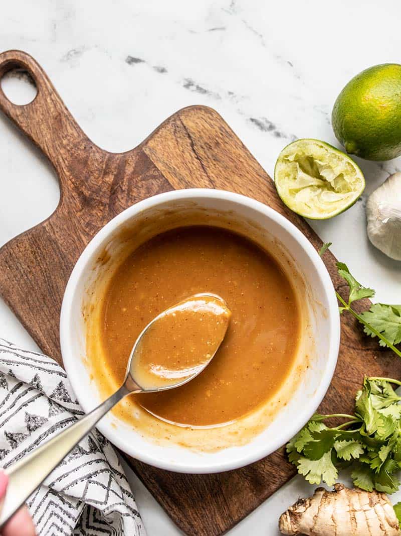 A spoon resting in a bowl of Peanut Lime Dressing, on a cutting board with limes, garlic, ginger, and cilantro all around.