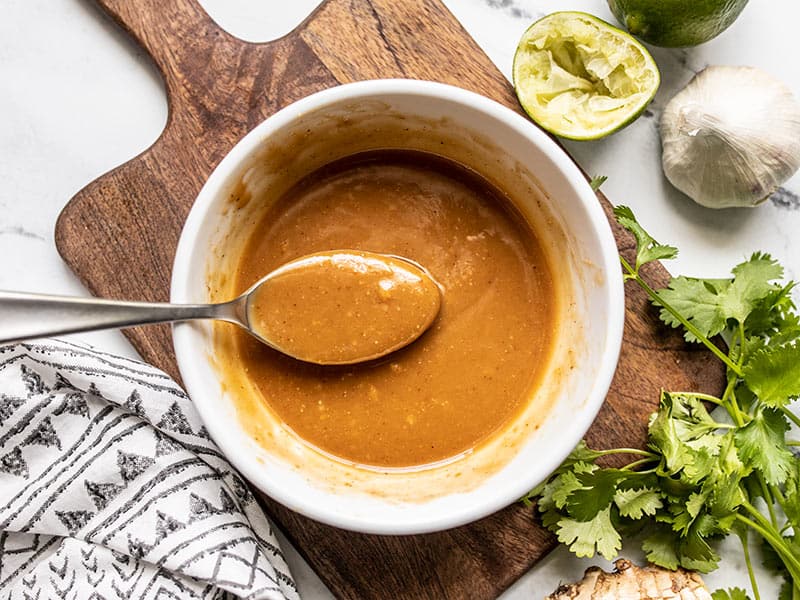 A spoon lifting some peanut lime dressing out of a bow, squeezed lime and cilantro on the side.