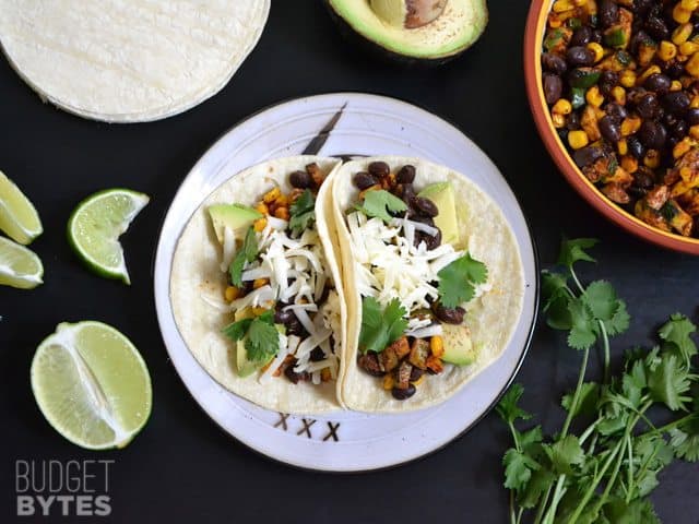 Top view of a plate of two Roasted Corn & Zucchini Tacos with extra toppings on the side 