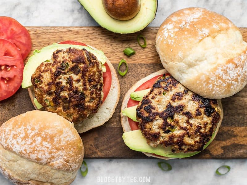 Two Green Chile Turkey Burgers on a wooden cutting board, being dressed. 