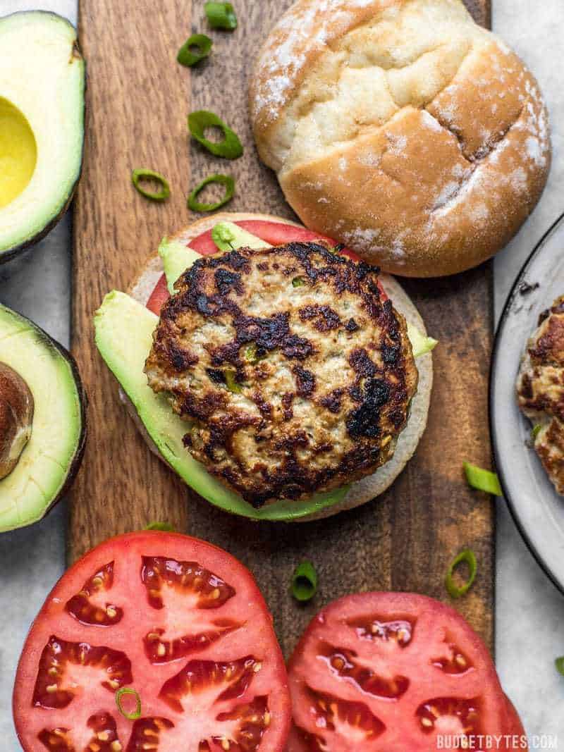 Green chile turkey burgers on a wooden cutting board next to tomato slices and avocado.