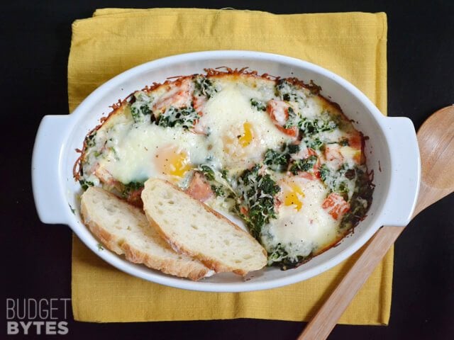 Baked Eggs with Spinach and Tomatoes in a dish sitting on a yellow napkin with a wooden spoon on the side 