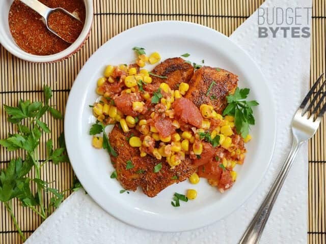 Top view of a plate of Blackened Tilapia with Corn salad on stop, small bowl of extra seasoning and fork on the side 