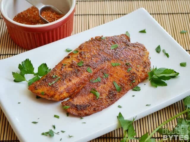 Blackened Tilapia filet on white plate garnished with parsley, small bowl of extra seasoning on the side 