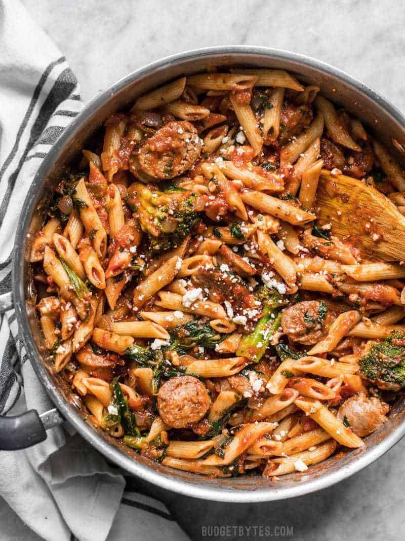 This super easy and satisfying, "sweep the kitchen" Penne Pasta with Sausage and Greens is an entire meal in one pot. Cook once, eat all week. A large pot full of Penne Pasta with Sausage and Greens with feta sprinkled over top and a wood spoon