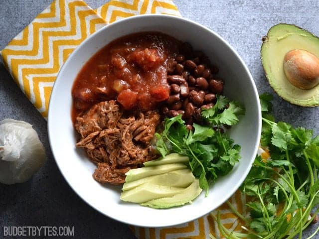 Top view of a bowl of southwest beef with salsa, cilantro, avocado and black beans sitting on a yellow chevron napkin with half an avocado and cilantro on the side 