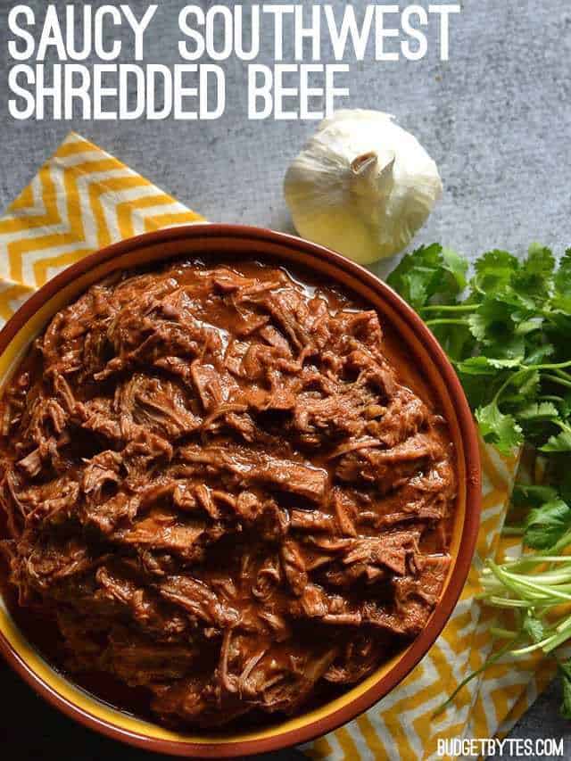 Top view of a bowl of Saucy Southwest Shredded Beef  sitting on a yellow chevron napkin with a clove of garlic and cilantro on the side