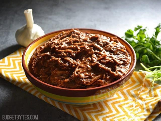 Side view of a bowl of Saucy Southwest Shredded Beef  sitting on a yellow chevron napkin with a clove of garlic and cilantro on the side