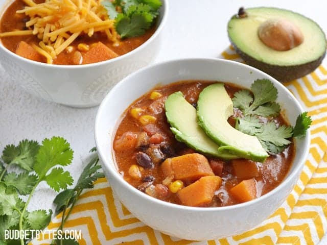 Side view of two bowls of Sweet Potato Tortilla Soup, one garnished with avocado and cilantro. Yellow chevron napkin, half an avocado and cilantro on the side for staging. 