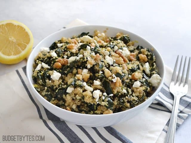 Side view of a bowl of Lemony Kale and Quinoa Salad sitting on a gray and white stripped napkin with a fork and half a lemon on the side 