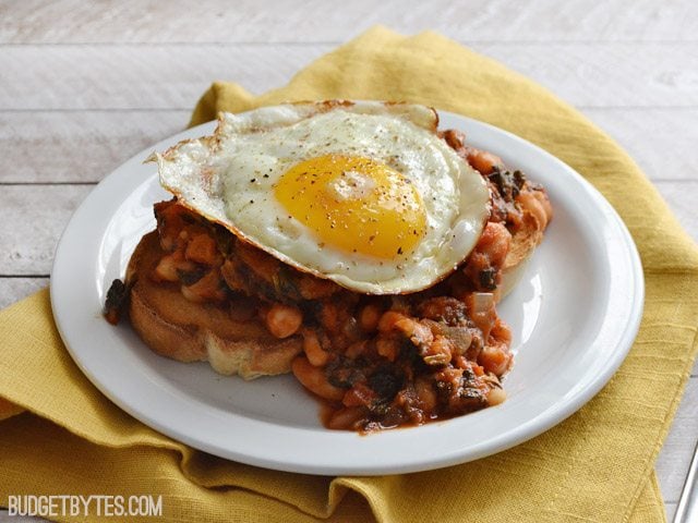 White Beans with Tomato and Sausage on a slice of bread with a fried egg on top, plate sitting on a yellow napkin 