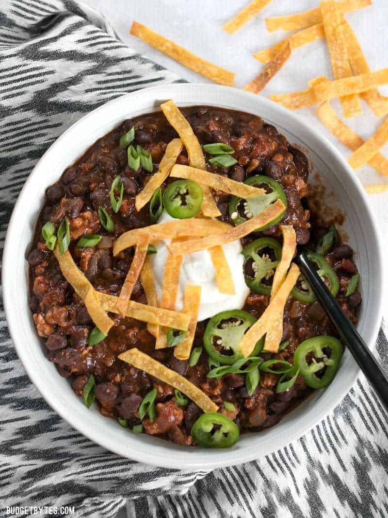 A big bowl of Weeknight Black Bean Chili with sour cream, jalapeño, and crispy tortilla strips.