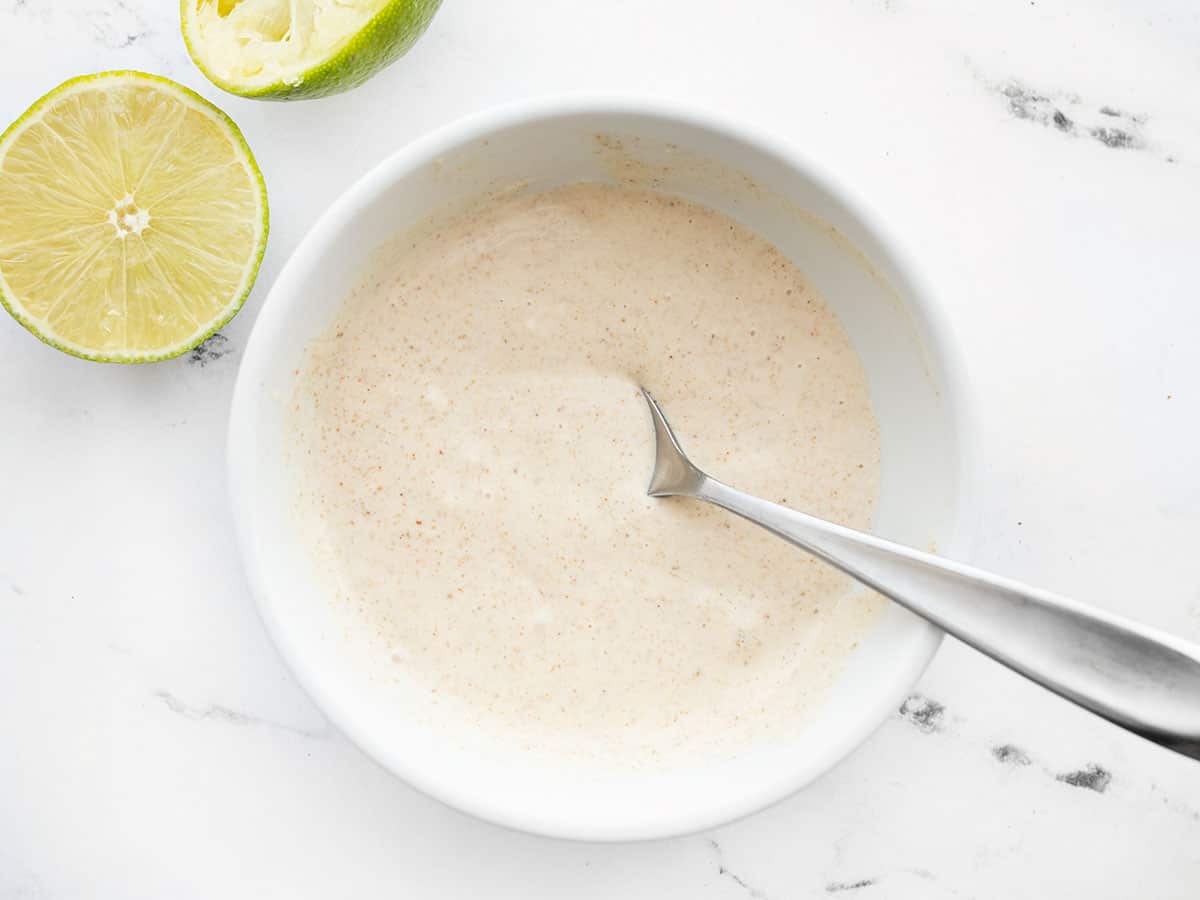 Overhead view of the cumin lime coleslaw dressing in a bowl with a spoon, cut limes on the side