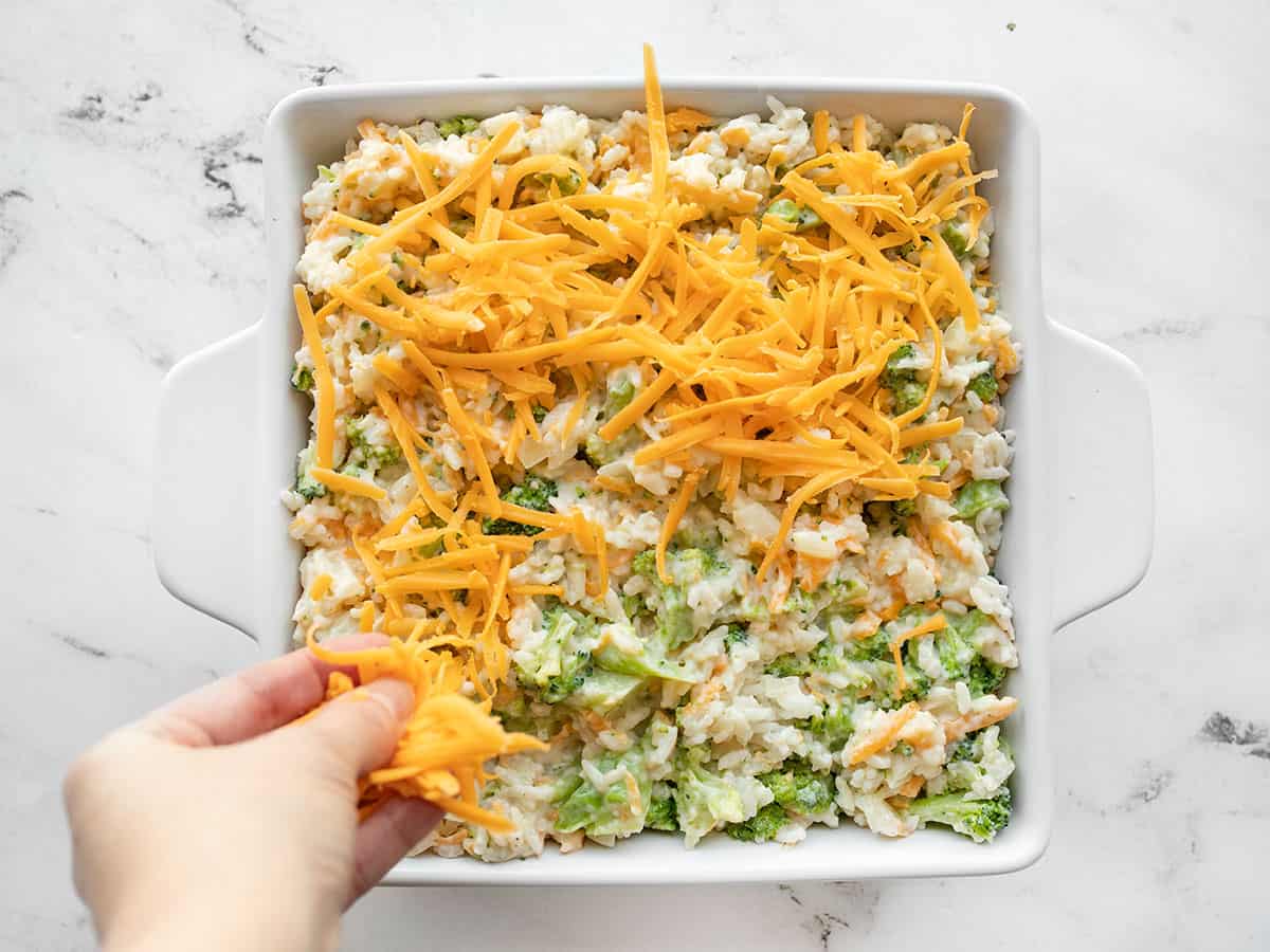 Broccoli rice mixture in a casserole dish being covered with shredded cheddar