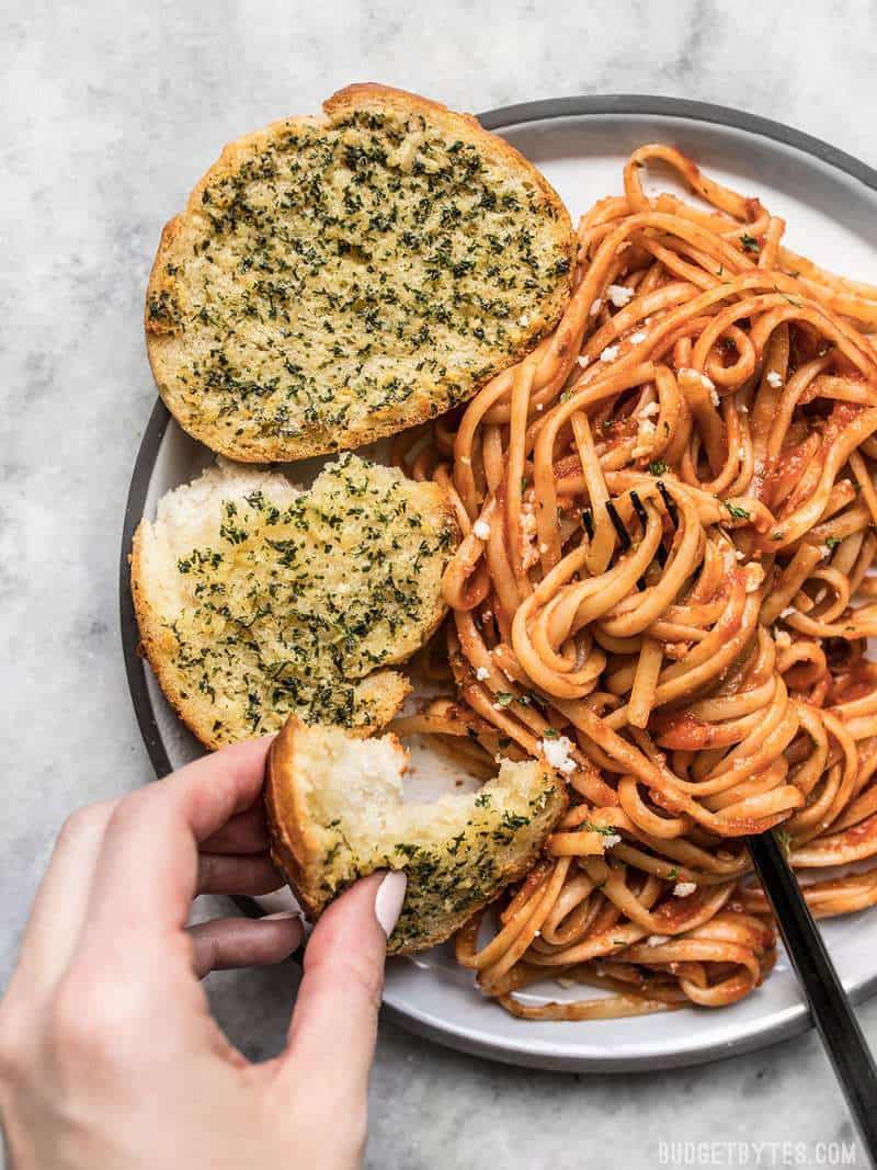 Two garlic bread slices on a plate of pasta with red sauce, a hand dipping a piece of bread in the sauce