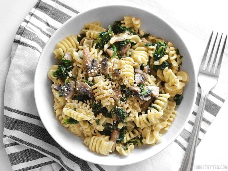 Top view of a bowl of Parmesan & Pepper Kale Pasta siting on a gray and white stripped napkin with a fork on the side