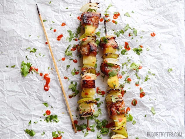 Two Pineapple Pork Kebabs garnished with cilantro and sriracha next to an empty skewer
