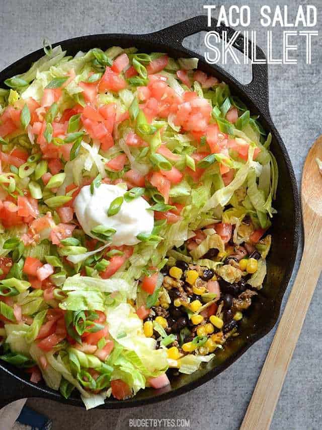 This entire taco salad is served right in the skillet used to cook the seasoned taco beef. Taco Salad Skillet is a fun, fast, and easy weeknight dinner. Budetbytes.com Overhead view of taco salad skillet with a portion scooped out, title text at the top.