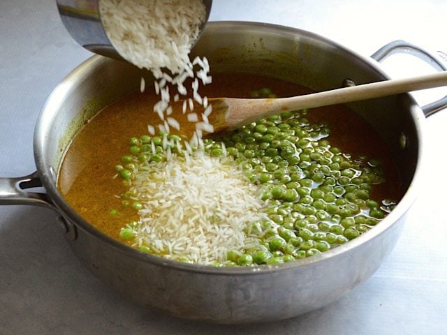 Rice being poured into the skillet with the broth and peas