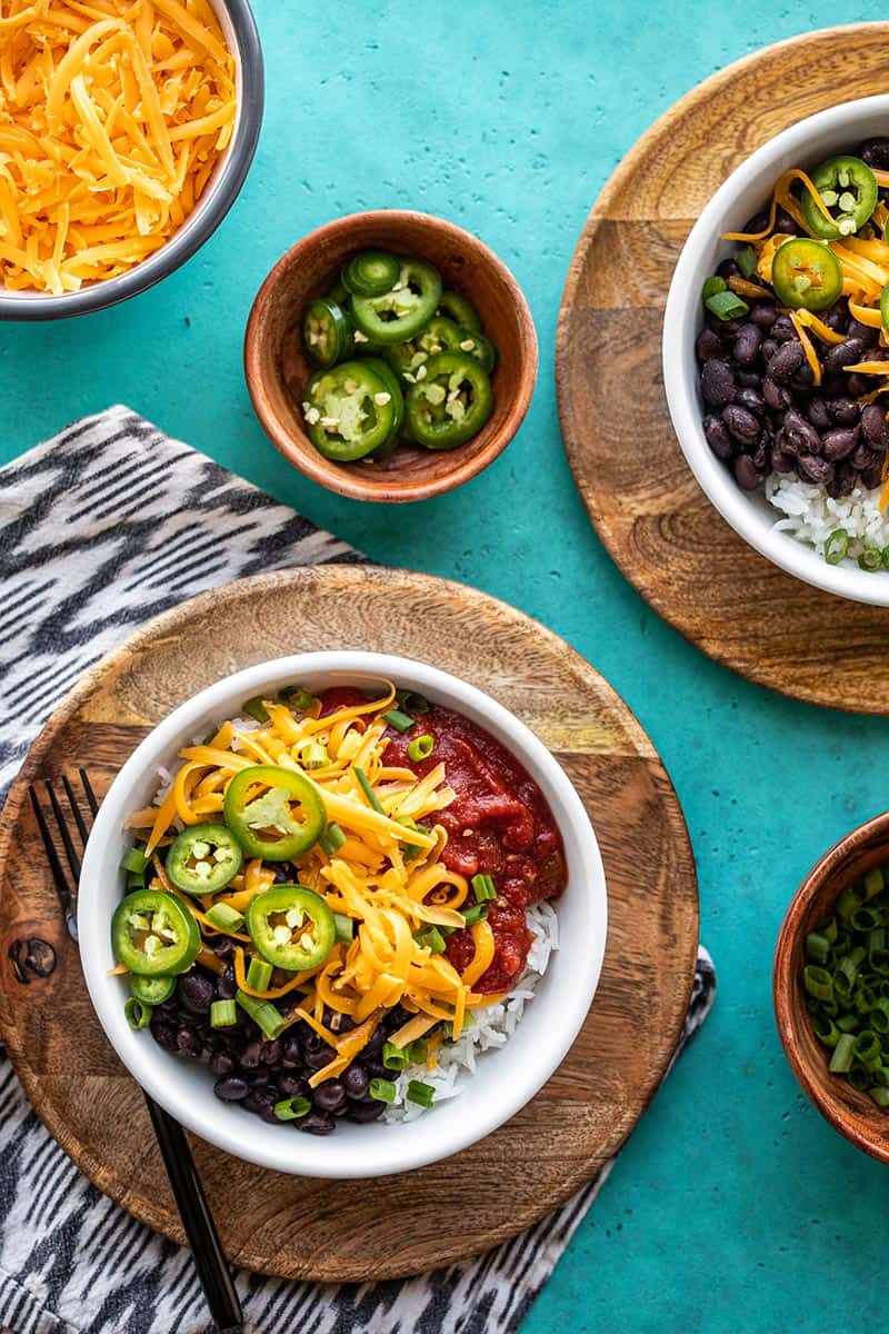 Two poor man's burrito bowls on wooden plates with bowls of toppings on the sides