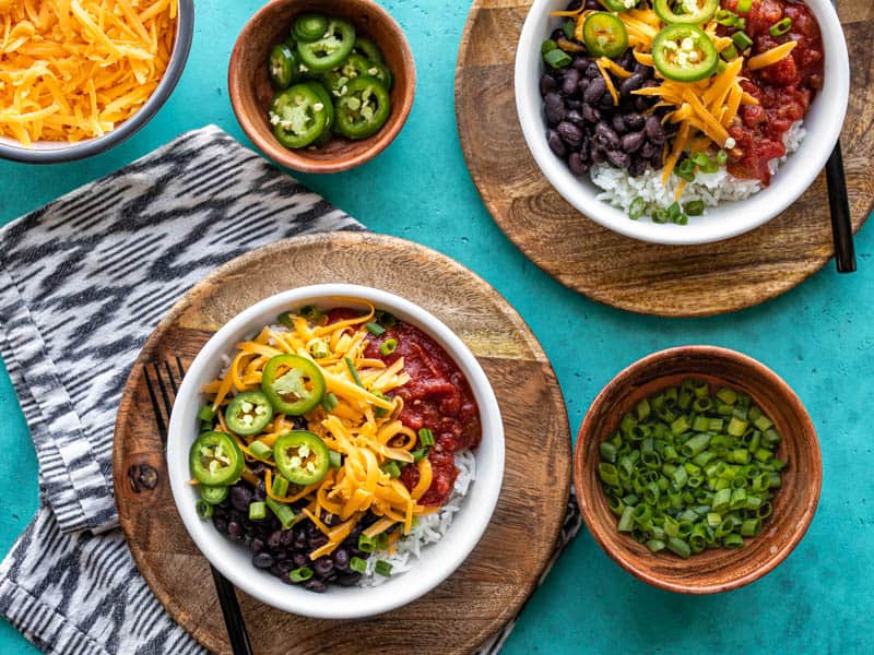 Two poor man's burrito bowls on wooden plates with bowls of toppings on the side