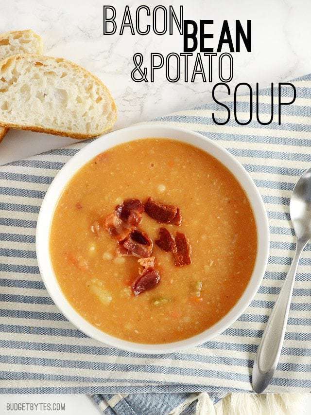 This hearty Bacon Bean and Potato Soup is extra thick with both beans and potato, plus enough bacon and vegetables to give each bowl plenty of flavor! Budetbytes.com Overhead view of a bowl of Bacon Bean and Potato Soup with bread on the side and title text at the top