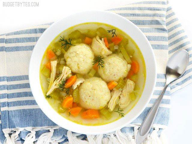 Top view of a bowl of Matzo Ball Soup sitting on a blue and white stripped napkin with a spoon on the side 