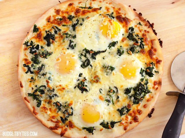 A whole Eggs Florentine Breakfast Pizza on a wooden cutting board, pizza slicer on the side.