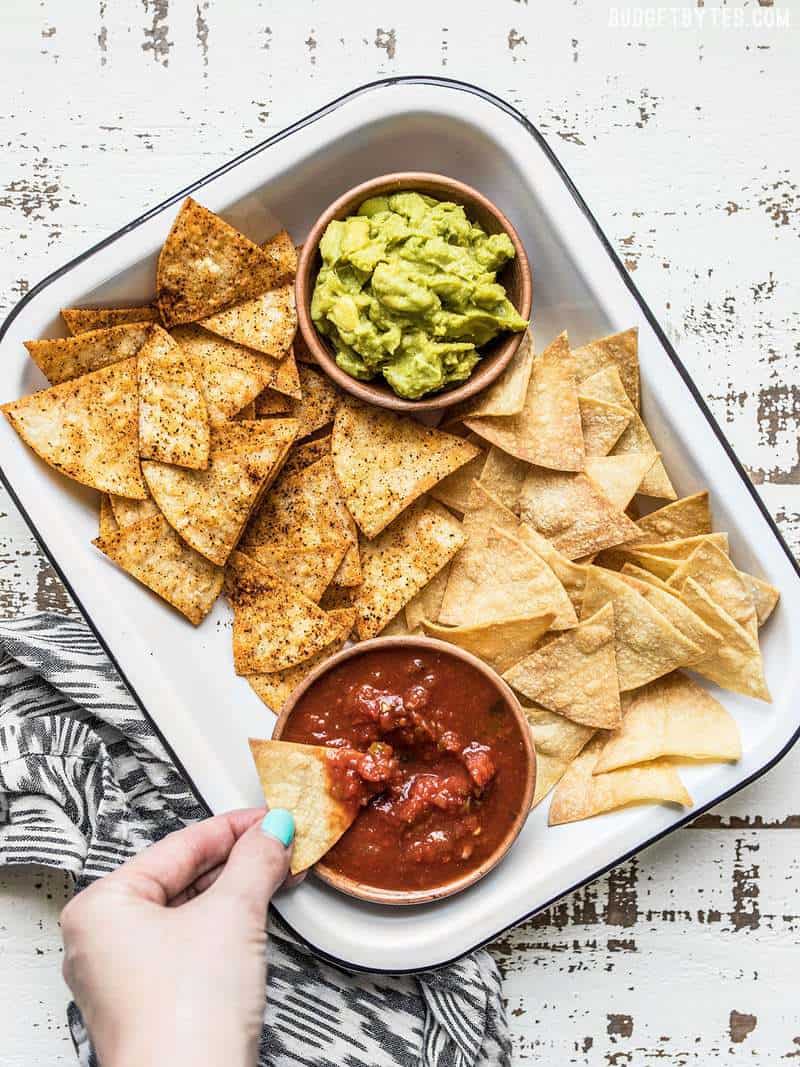 Homemade Baked Tortilla Chips in a baking dish with small bowls of salsa and guacamole, a hand dipping one chip
