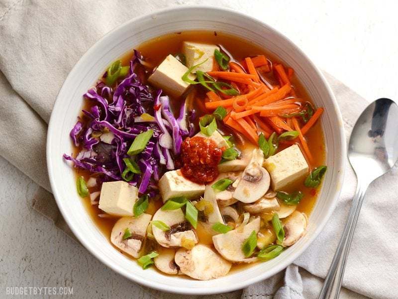 Overhead view of a bowl full of hot and sour soup with vegetables and cubed tofu, chili garlic sauce in the center of the bowl