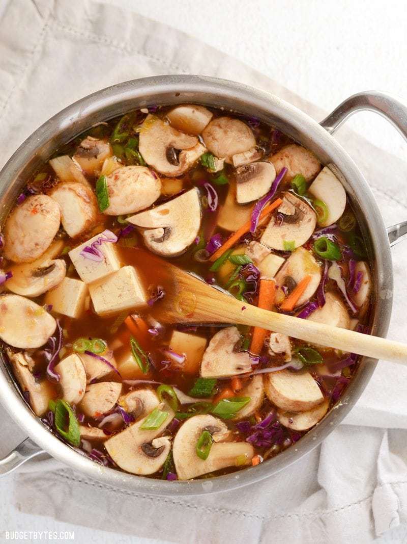 Overhead view of a pot full of hot and sour soup with colorful vegetables and cubes of tofu. A wooden spoon in the center.